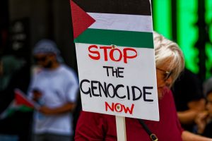 Woman checking her phone and holding a sign "stop the genocide".
